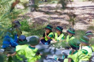 Little Scholars kids wearing matching green sun hats and high-visibility yellow vests sit in a circle on a grey groundsheet in a wooded area, listening intently to an adult educator.