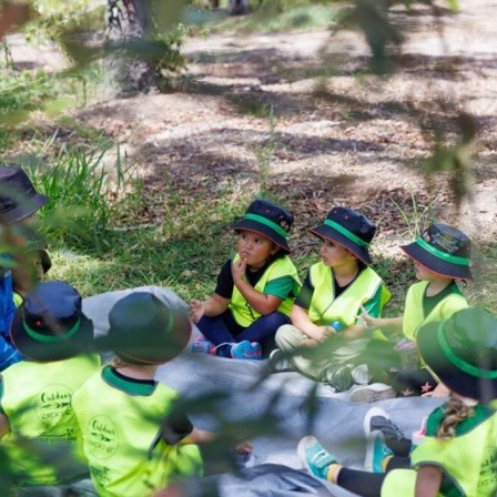 Little Scholars kids wearing matching green sun hats and high-visibility yellow vests sit in a circle on a grey groundsheet in a wooded area, listening intently to an adult educator.