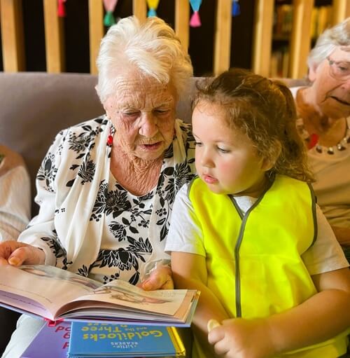 Elderly And Young Child Reading A Storybook Together During A Little Scholars Intergenerational Program