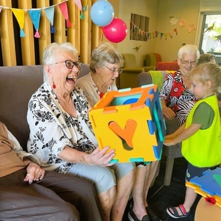 Elderly Women Laughing Happily While Playing With A Large Shape Sorter Puzzle Cube With A Young Little Scholars Student