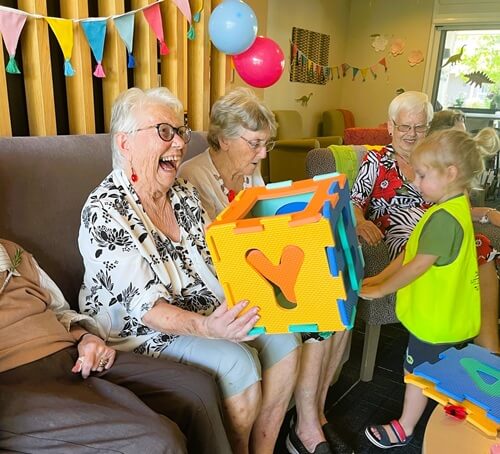 Elderly Women Laughing Happily While Playing With A Large Shape Sorter Puzzle Cube With A Young Little Scholars Student