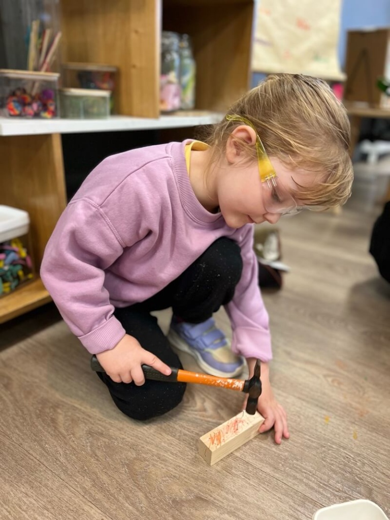 A young girl with safety glasses is kneeling on a wooden floor, focused intently on hammering a small nail into a block of wood. She is wearing a light purple sweater and black pants. This demonstrates practical life skills and concentration during a woodworking activity.