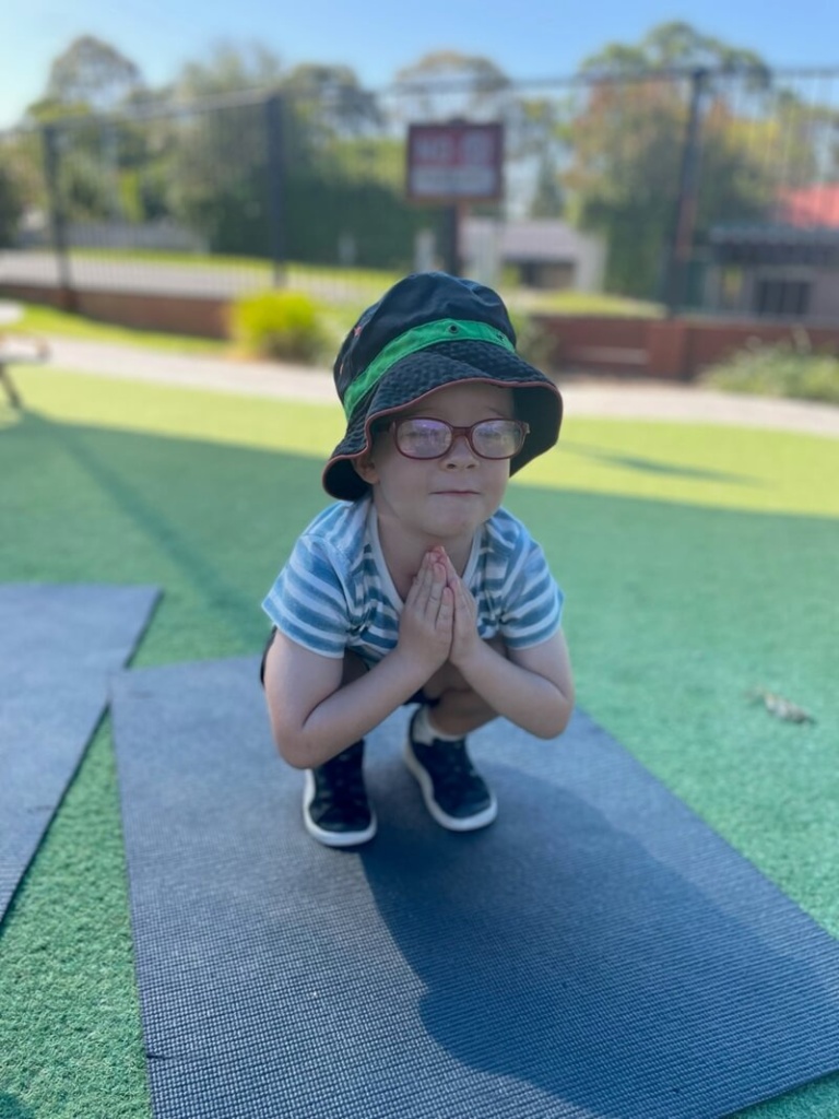 A Young Boy Wearing A Striped Shirt, Black Bucket Hat, And Glasses Is Practicing Yoga Or Mindfulness At Little Scholars School Of Early Learning