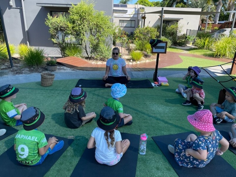 An outdoor view of a group of preschool students sitting on yoga mats in a circle on green artificial turf, participating in a mindfulness or yoga class. A female educator in a white smiley-face t-shirt is leading the activity, seated cross-legged on a mat in the center. The children are wearing sun hats and are engaging in the lesson.