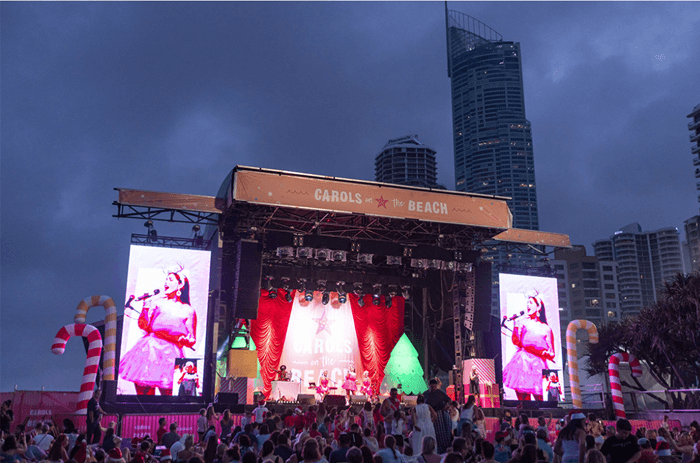 Santa, the Grinch, and festive elves perform on a brightly lit stage in front of a massive crowd at a large Christmas carols event on the beach