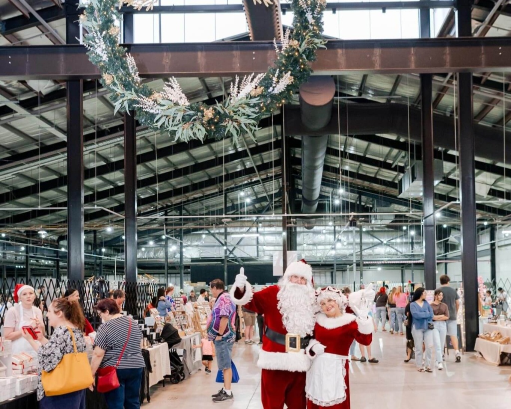 Santa and Mrs. Claus pose for a photo with a large festive wreath hanging overhead inside a large, bustling indoor market venue on the Gold Coast.