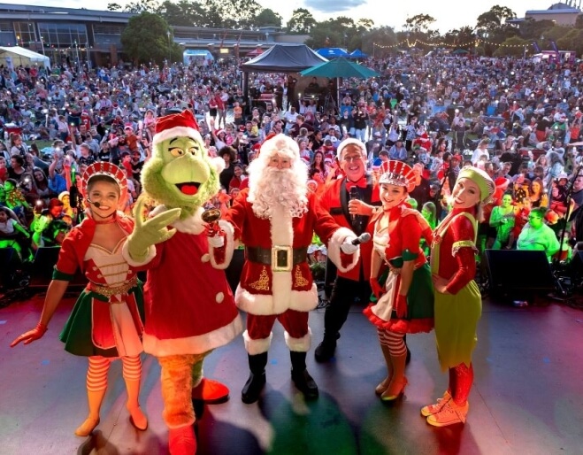 Santa, the Grinch, and festive elves perform on a brightly lit stage in front of a massive crowd at a large Christmas carols event on the Gold Coast.