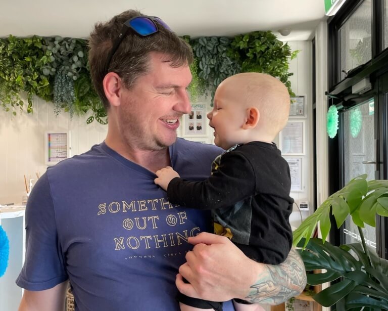 A loving father wearing a blue shirt holds his baby son, with both smiling at each other in a joyful moment inside the Little Scholars early learning center, highlighting the importance of family and bonding.