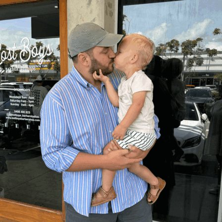 A father wearing a blue striped shirt and baseball cap holds his toddler child, who is giving him a sweet kiss on the mouth outside a barbershop, emphasizing the loving connection between Little Scholars families.