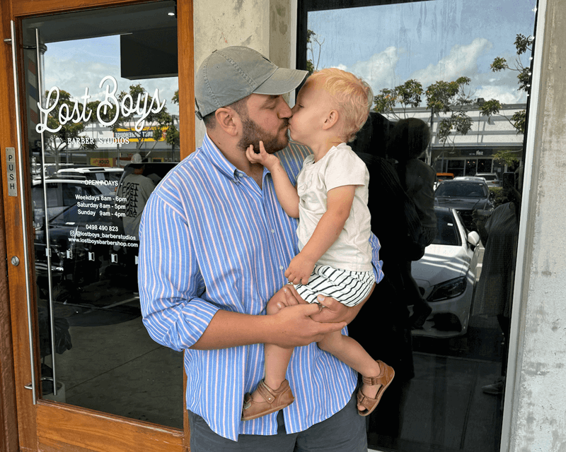 A father wearing a blue striped shirt and baseball cap holds his toddler child, who is giving him a sweet kiss on the mouth outside a barbershop, emphasizing the loving connection between Little Scholars families.