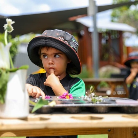 A curious preschool child wearing a sun-safe hat concentrates on sorting natural materials (flowers and leaves) on an outdoor wooden table, highlighting sensory and nature exploration at the Little Scholars campus.