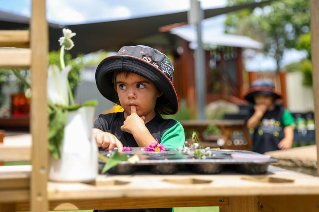 A curious preschool child wearing a sun-safe hat concentrates on sorting natural materials (flowers and leaves) on an outdoor wooden table, highlighting sensory and nature exploration at the Little Scholars campus.