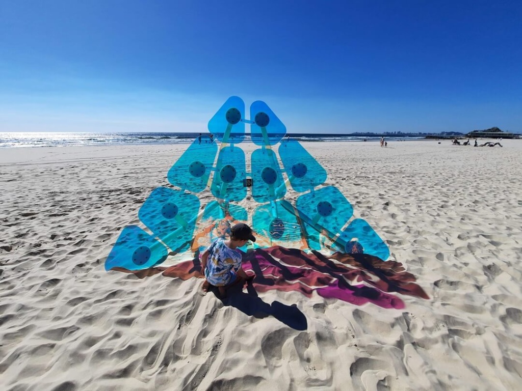Child interacting with a large blue pyramid-shaped art installation on a sunny, white sand beach with the ocean and blue sky in the background.
