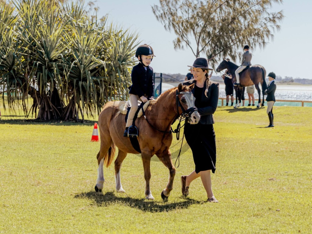 A child wearing a helmet riding a pony led by an instructor on a sunny grassy field by the water, possibly during an equestrian event.