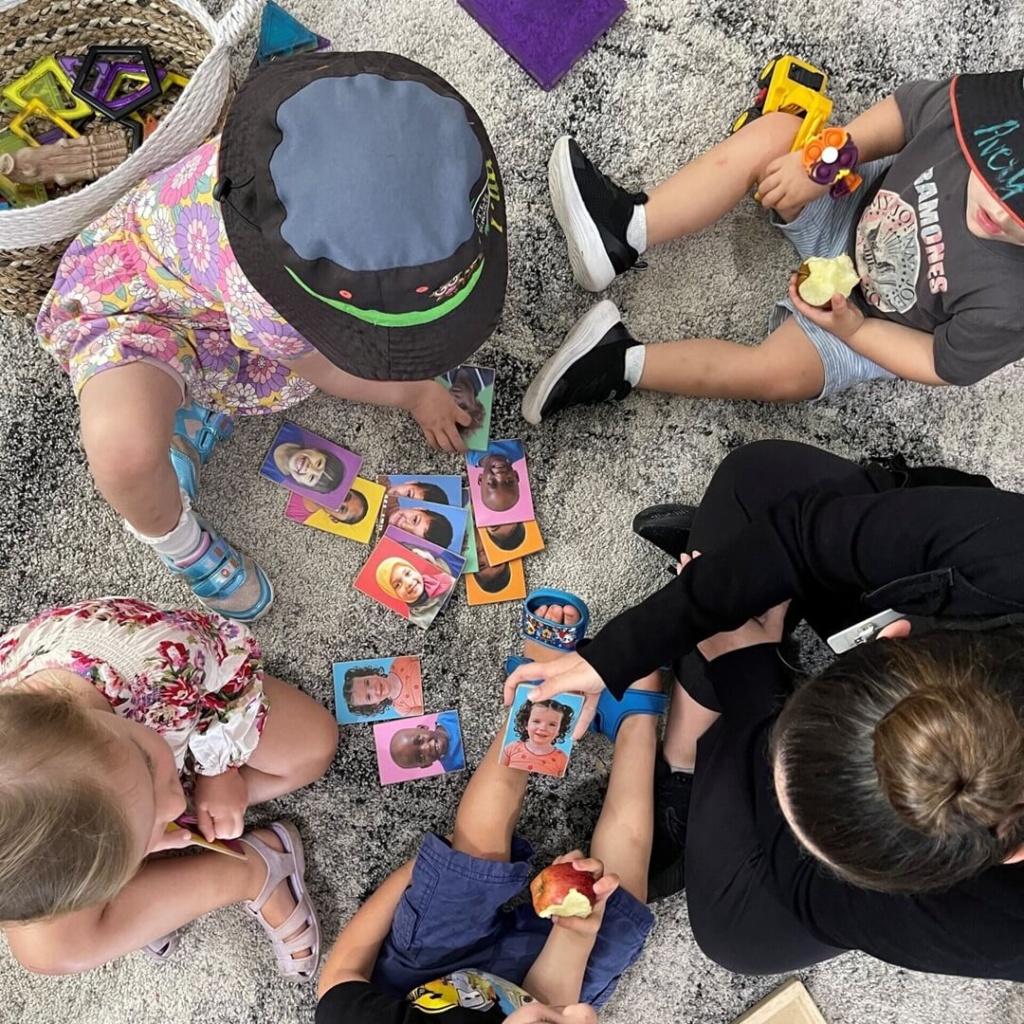 Overhead view of a Little Scholars educator and four children sitting on a rug, engaging with diverse emotion flashcards for social and emotional learning and awareness