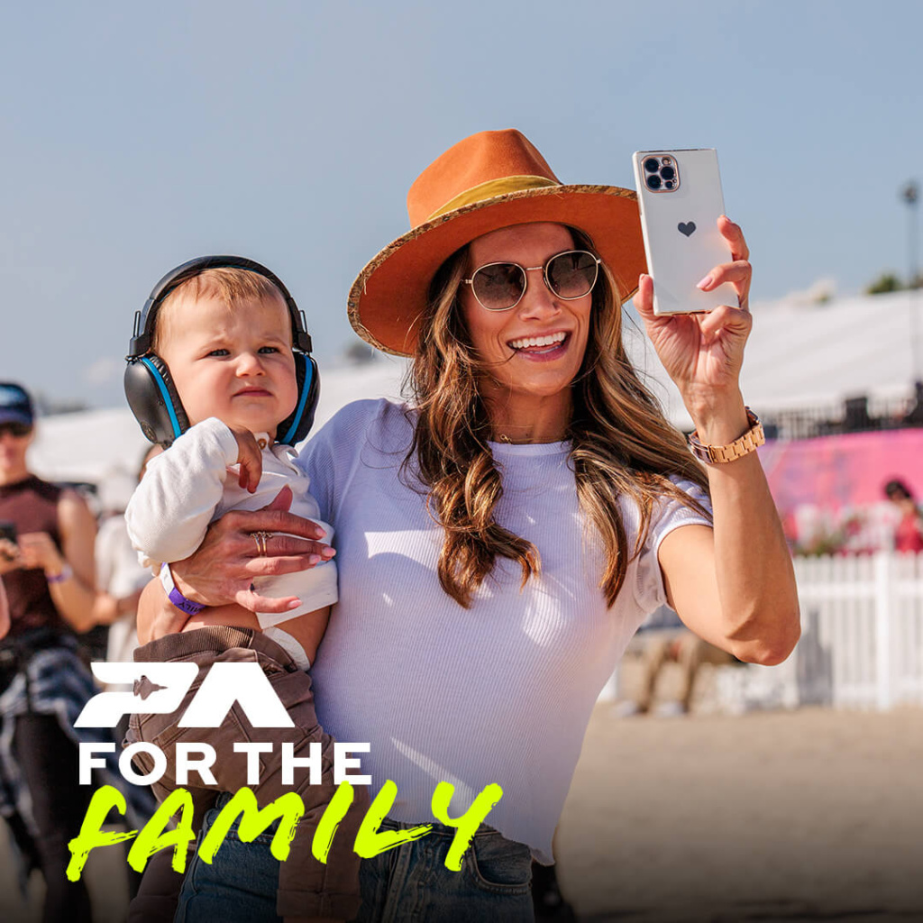 Happy mother taking a selfie on her phone while holding a baby wearing noise-cancelling headphones at a sunny outdoor event.