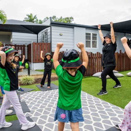 Children and educators at Little Scholars daycare doing yoga on mats, wearing green uniforms and hats, in an outdoor Gold Coast childcare setting with a modern building and greenery.