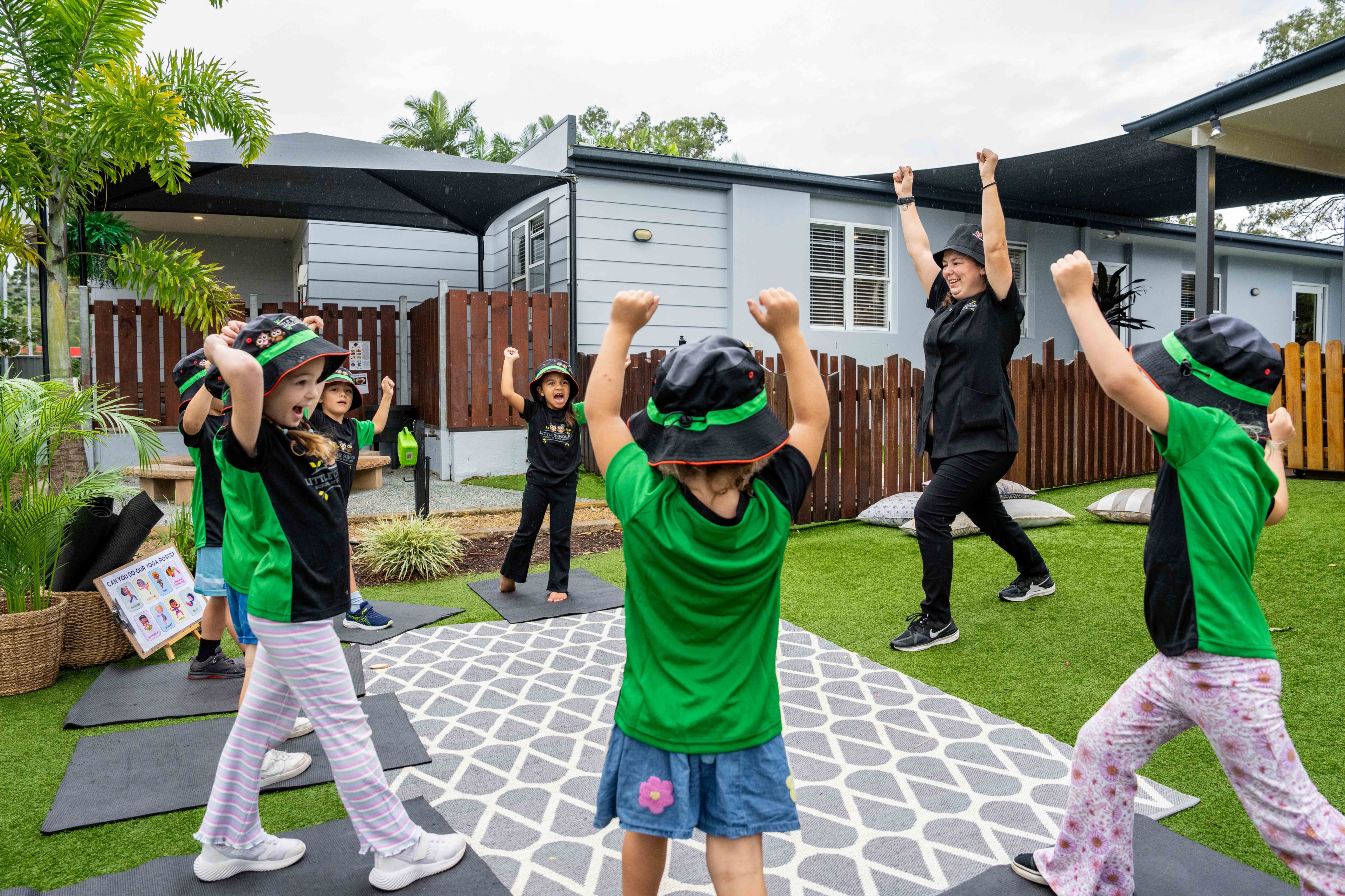 Children and educators at Little Scholars daycare doing yoga on mats, wearing green uniforms and hats, in an outdoor Gold Coast childcare setting with a modern building and greenery.