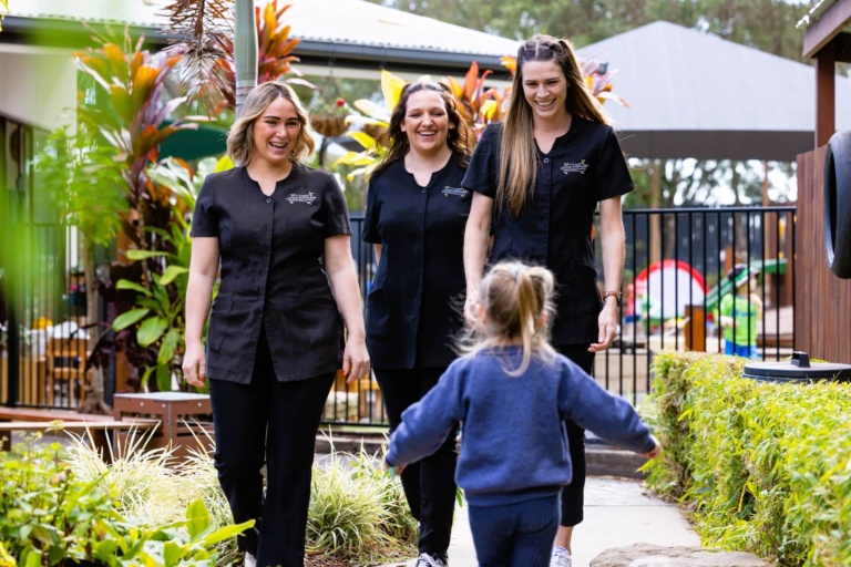 Childcare educators at Little Scholars greet a young child in an outdoor play area.