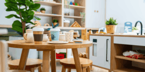 Bright, modern Little Scholars classroom interior with natural wooden tables and chairs, shelves stocked with resources, and a dramatic play kitchen area featuring toy coffee cups