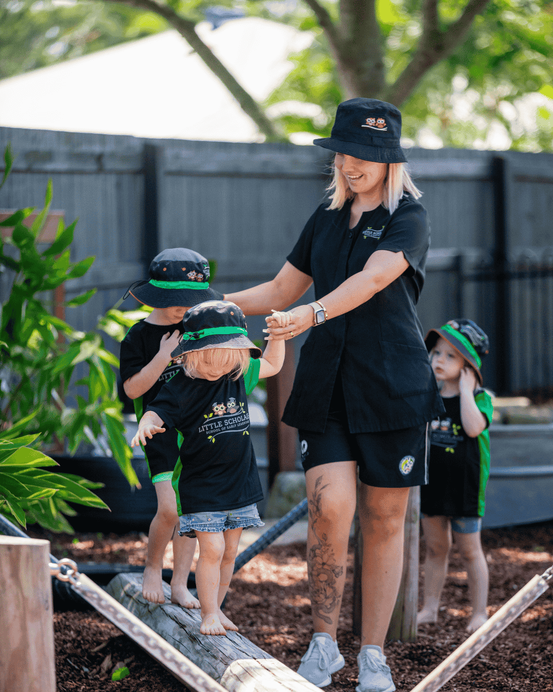 An early childhood educator at Little Scholars safely supervising two toddlers walking on a low log balance beam in an outdoor play area