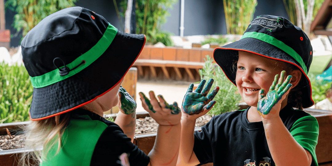 Two happy children from the Little Scholars Senior Kindy program with hands covered in green finger paint, engaging in play-based learning outdoors