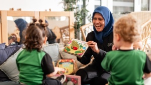 Smiling female early childhood educator wearing a blue hijab reading an interactive pop-up book to two toddlers during story time at Little Scholars