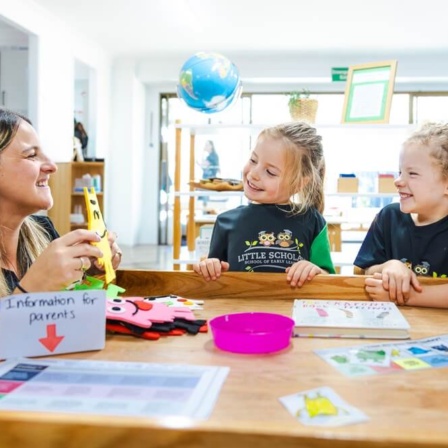 A high-quality photo of a smiling female educator with long, dark hair sitting at a wooden table and warmly interacting with two young, blonde children (a girl and a boy) wearing green and black Little Scholars uniforms. The three are focused on an object the educator is holding. Various learning materials, including a book, flashcards, a pink bowl, and a sign that reads "Information for parents," are spread across the table. A globe hangs in the background of the bright, well-lit classroom.
