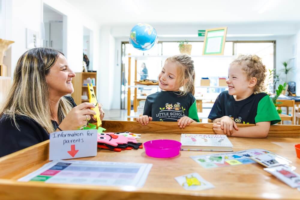 A high-quality photo of a smiling female educator with long, dark hair sitting at a wooden table and warmly interacting with two young, blonde children (a girl and a boy) wearing green and black Little Scholars uniforms. The three are focused on an object the educator is holding. Various learning materials, including a book, flashcards, a pink bowl, and a sign that reads "Information for parents," are spread across the table. A globe hangs in the background of the bright, well-lit classroom.
