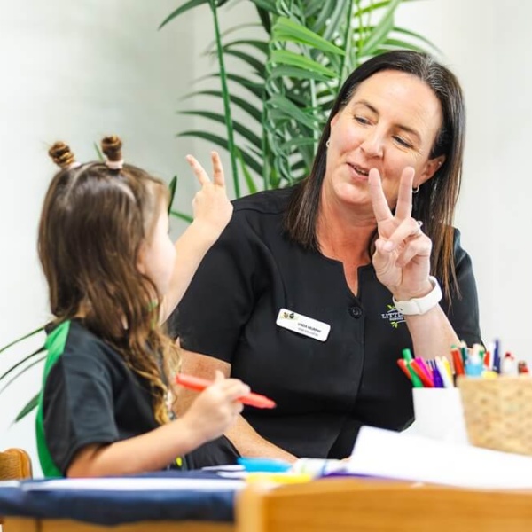 A close-up, brightly lit photo showing a smiling female educator wearing a black Little Scholars uniform and a white wristwatch, making a peace sign (two fingers up) while looking at a young girl. The girl, who has her hair tied up in playful knots, is sitting opposite her at a table, also holding up a peace sign. Both are engaged in an activity, and the girl holds a red pencil. A woven basket of colored pencils sits on the table between them, and a green potted plant is visible in the background.
