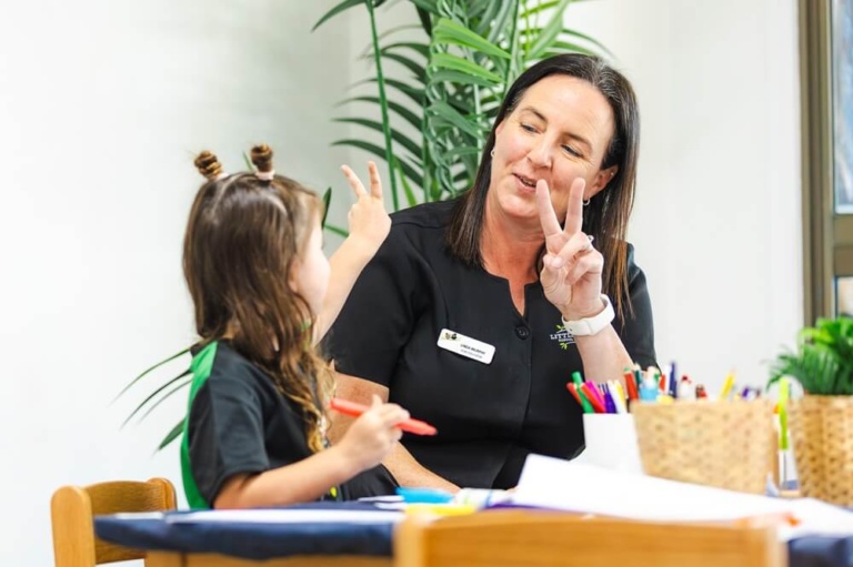 A close-up, brightly lit photo showing a smiling female educator wearing a black Little Scholars uniform and a white wristwatch, making a peace sign (two fingers up) while looking at a young girl. The girl, who has her hair tied up in playful knots, is sitting opposite her at a table, also holding up a peace sign. Both are engaged in an activity, and the girl holds a red pencil. A woven basket of colored pencils sits on the table between them, and a green potted plant is visible in the background.