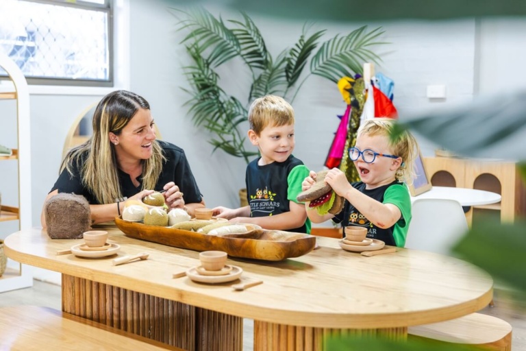 A female childcare educator sitting with two young boys at a wooden table engaging in sensory play with toy sandwiches at Little Scholars.