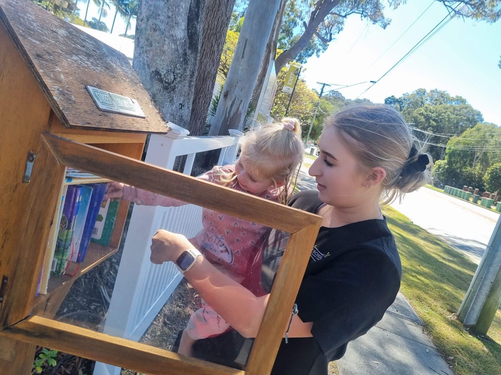Little Scholars Educator Holds A Young Child As They Look Through A Wooden Street Library Mail Box Filled With Books Outdoors