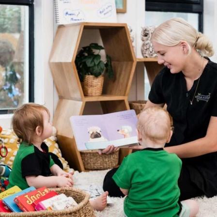 Two toddlers sit attentively on the floor listening to a Little Scholars educator reading a colorful picture book during story time, emphasizing early literacy and social-emotional development in the preschool environment.